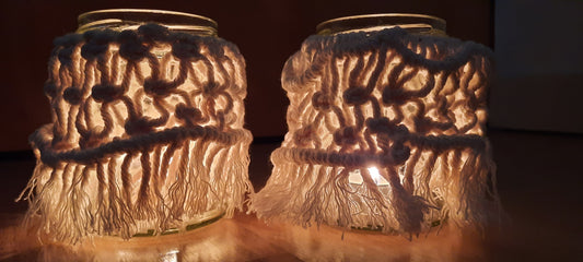 Two glass jars with woven covers and candles inside on a dark background