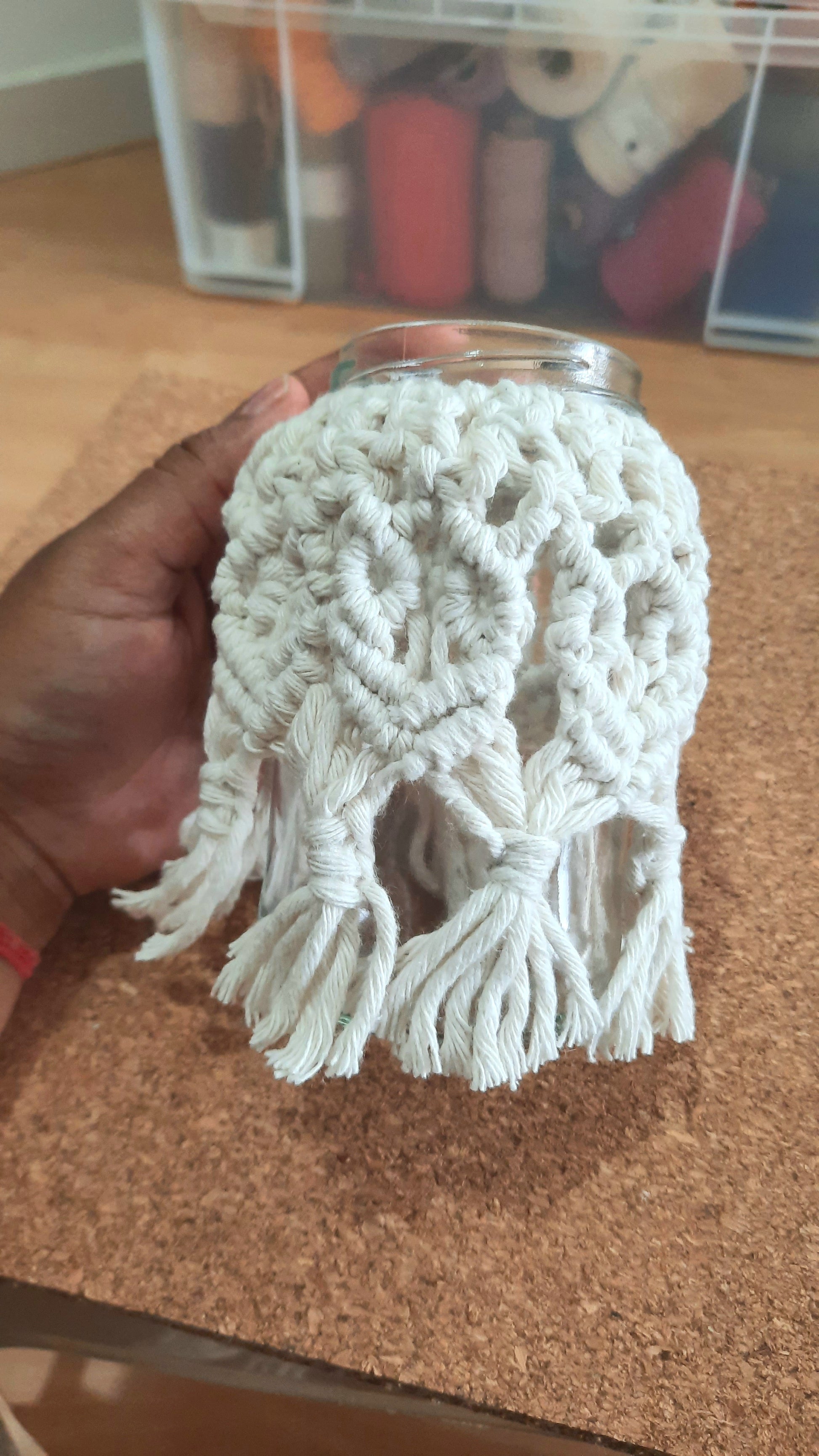 Hand holding a white crocheted doily over a glass jar on a cork surface.