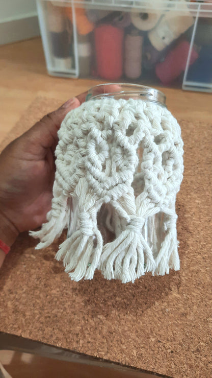 Hand holding a white crocheted doily over a glass jar on a cork surface.
